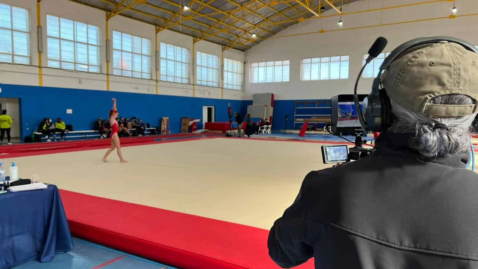 Operador de cámara con auriculares de intercom grabando un ejercicio de gimnasia desde el borde del tapiz, en un pabellón municipal.