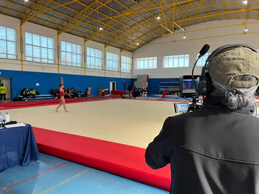 Operador de cámara con auriculares de intercom grabando un ejercicio de gimnasia desde el borde del tapiz, en un pabellón municipal.