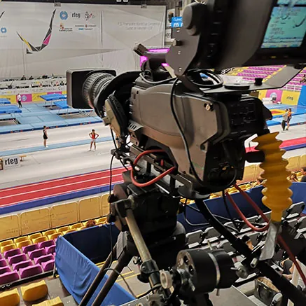 Cámara de televisión sobre trípode en la grada de un pabellón, enfocando la pista de competición antes de que llegue el público.