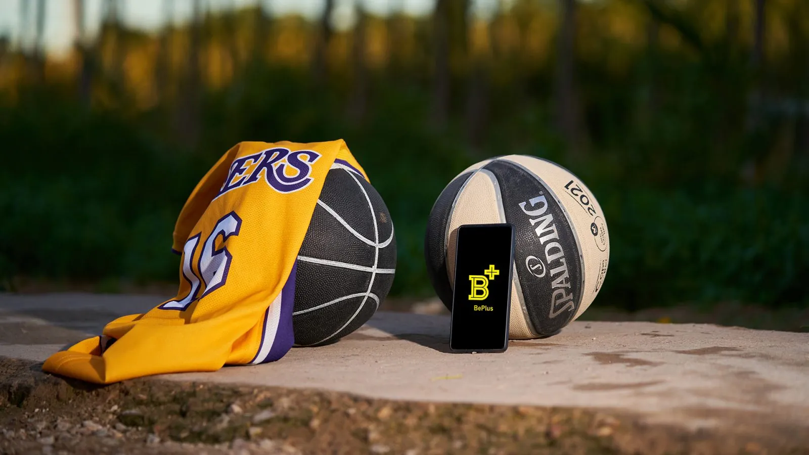 Balón de baloncesto y camiseta junto a un teléfono móvil mostrando la aplicación BePlus, sobre una pista exterior al atardecer.