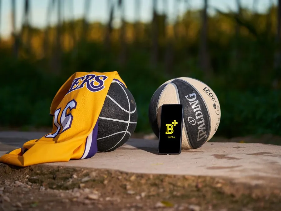 Balón de baloncesto y camiseta junto a un teléfono móvil mostrando la aplicación BePlus, sobre una pista exterior al atardecer.